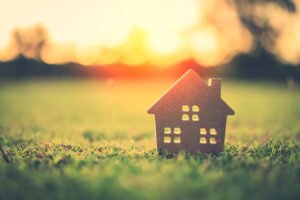 A small cardboard cut-out of a house placed on grass, with a warm sunset glowing in the background
